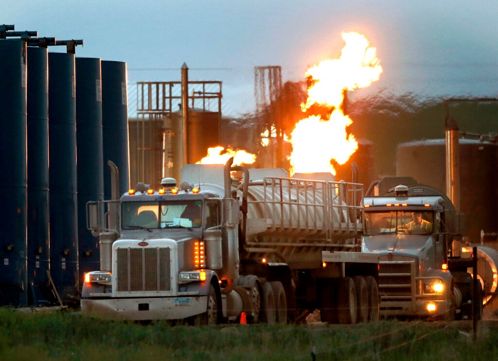 Two semi-trucks sit next to industrial tanks at a fracking pad as large orange flames and vapors shoot up behind them.