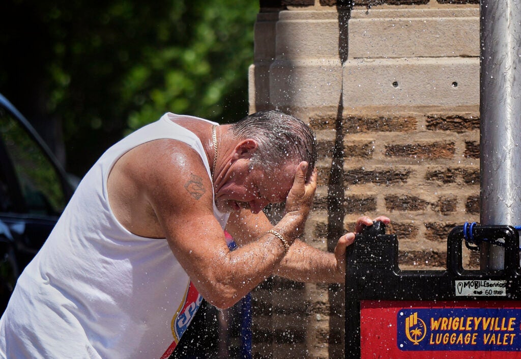 A man in a white tank top leans into a spray of water while holding his head.