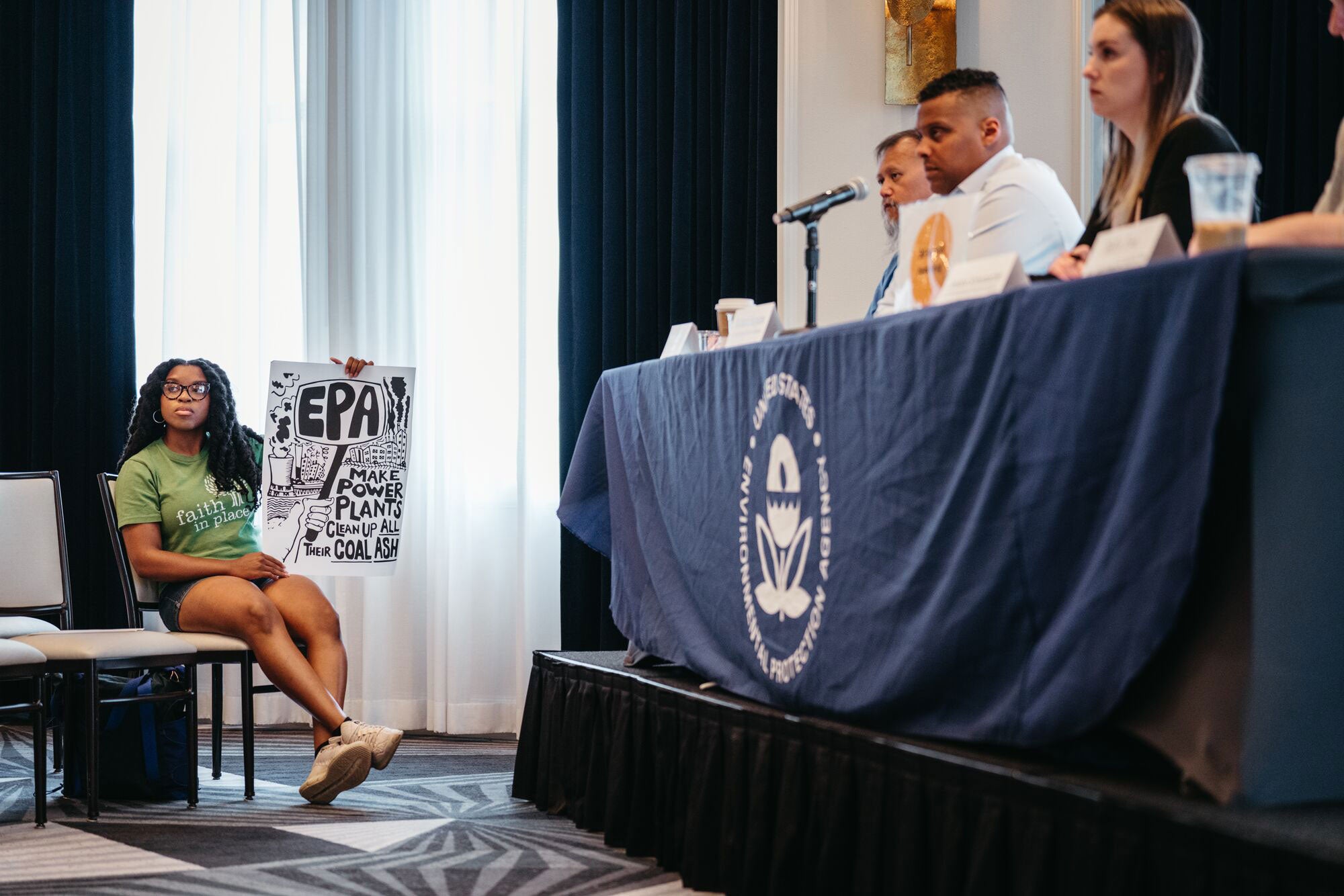 A panel of EPA officials listen as members of the public give testimony at the hearing, as a changemaker seated in the chair nearest to the panel holds a sign that reads, "EPA: Make Power Plants Clean Up All Their Coal Ash."