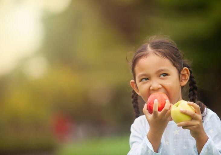 A young girl holds two apples in her hands. A red apple in one hand that she is taking a bite out of and a yellow/green apple in her other hand. A blurred green background.