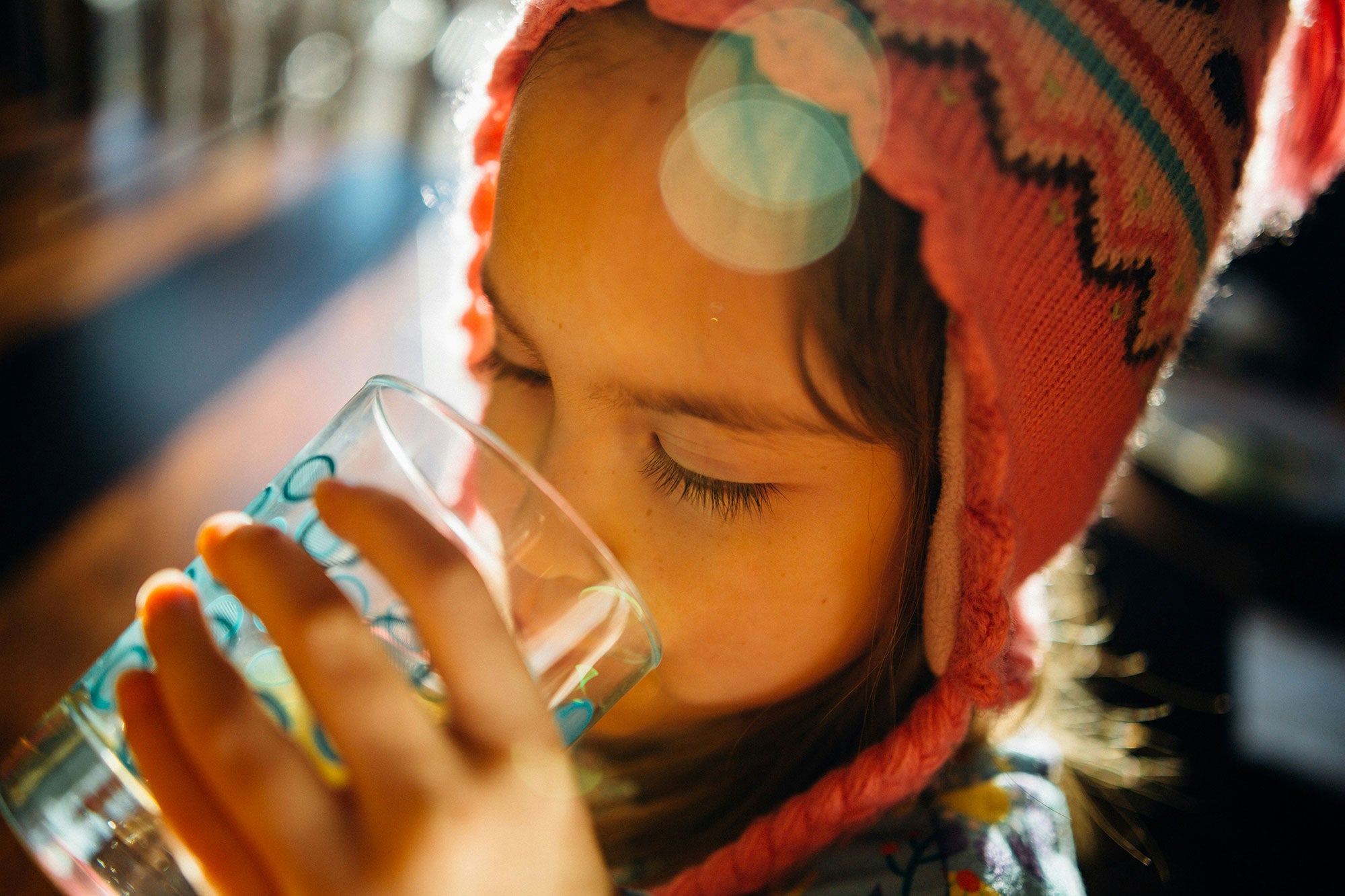 A top-down, close-up view of young child wearing a brightly colored knit cap, drinking a glass of water, backlit by the sun.