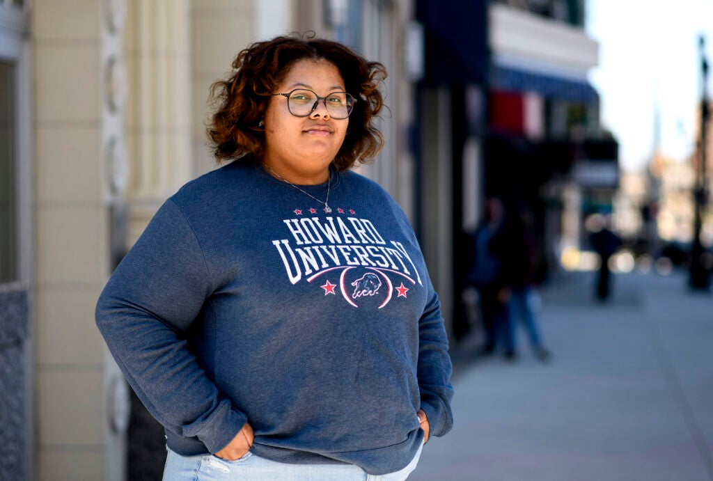 A woman stand on the sidewalk of a city downtown looking directly into the camera with her hands in her pocket. Her sweater reads "Howard University"