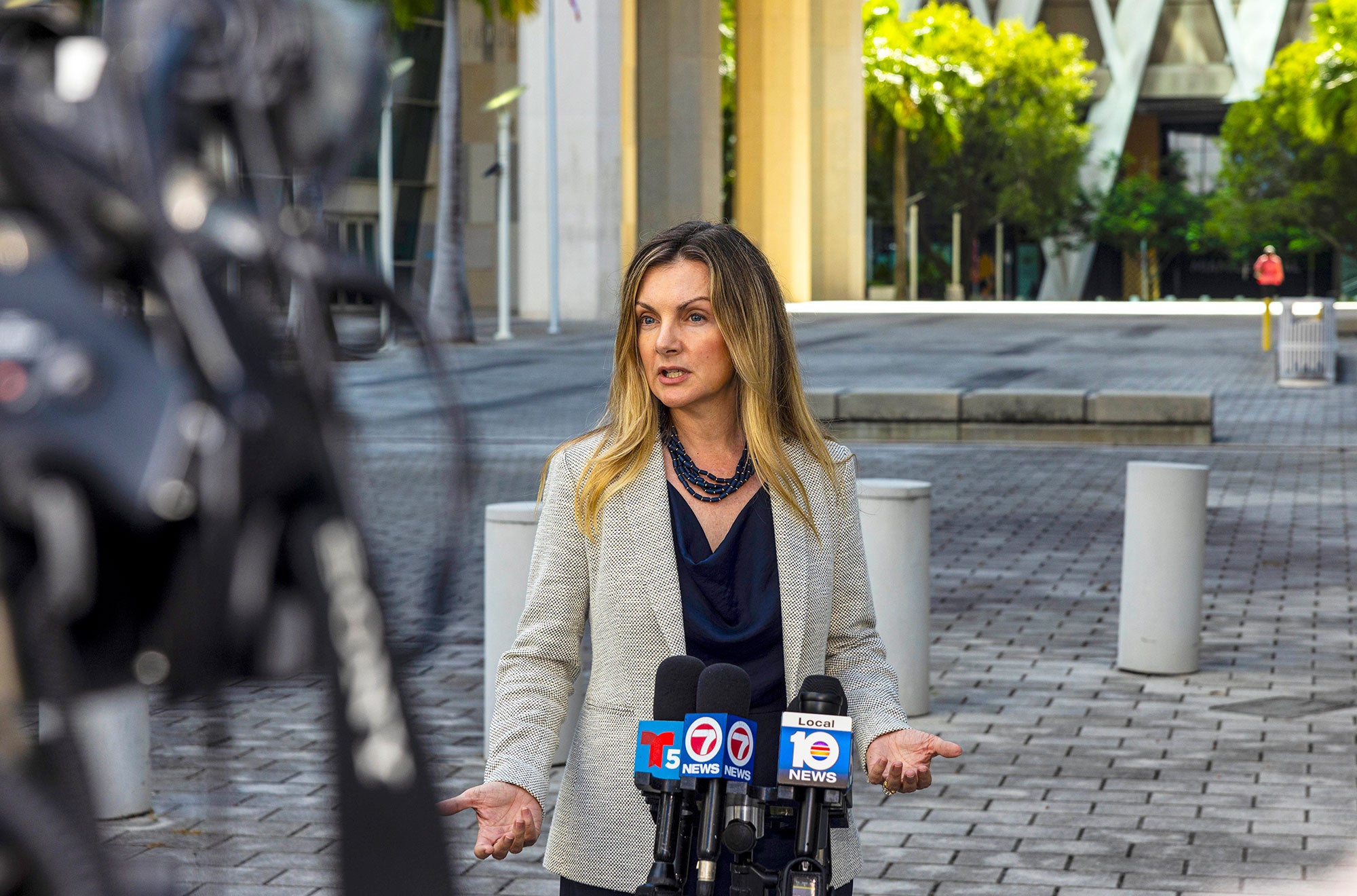 A woman wearing a light grey blazer and dark blue top speaks with her hands outstretched, in front of TV station microphones, while standing outdoors in the courtyard of a building. A TV crew camera is visible to the side.