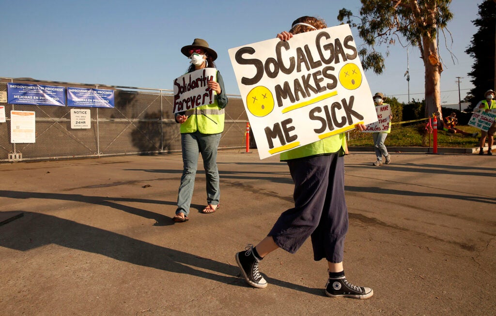 People hold signs in front of a gated facility. One says "SoCalGas makes me sick"