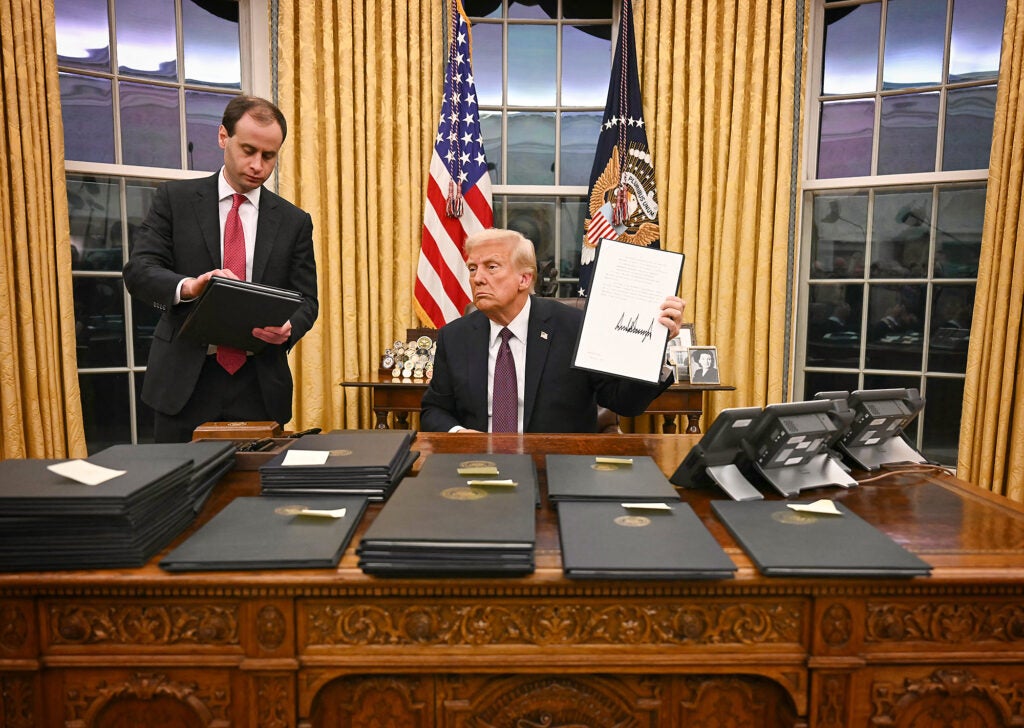 Donald Trump holds up a signed document while seated at his desk in the Oval Office, which is covered in stacks of document binders. He has a serious expression on his face and a man is standing next to him holding more document binders.