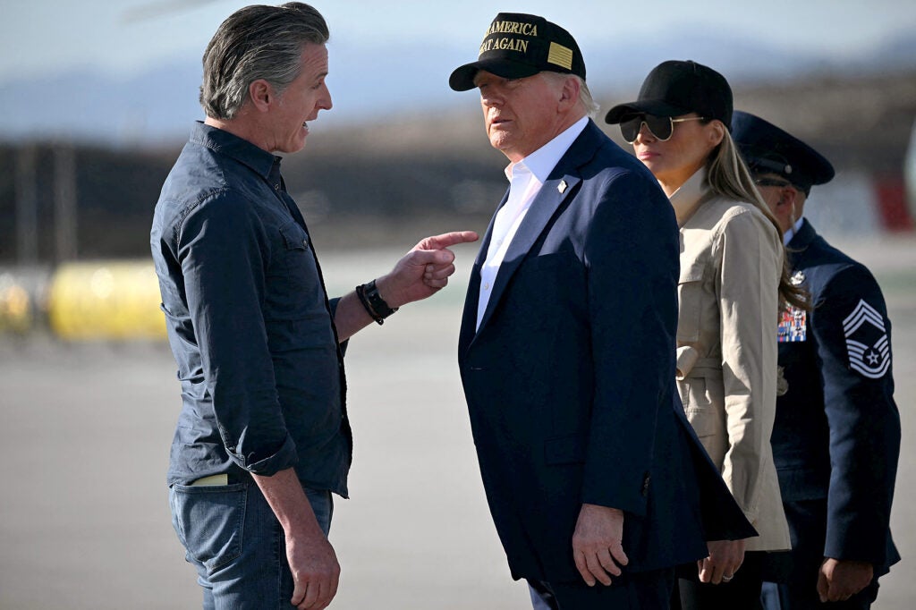 Gavin Newsom, wearing jeans and a blue collard shirt is pointing a finger at Donald Trump's chest while speaking to him with a serious expression. Trump, wearing a blue suit jacket and no tie and a black baseball hat, is looking at him.