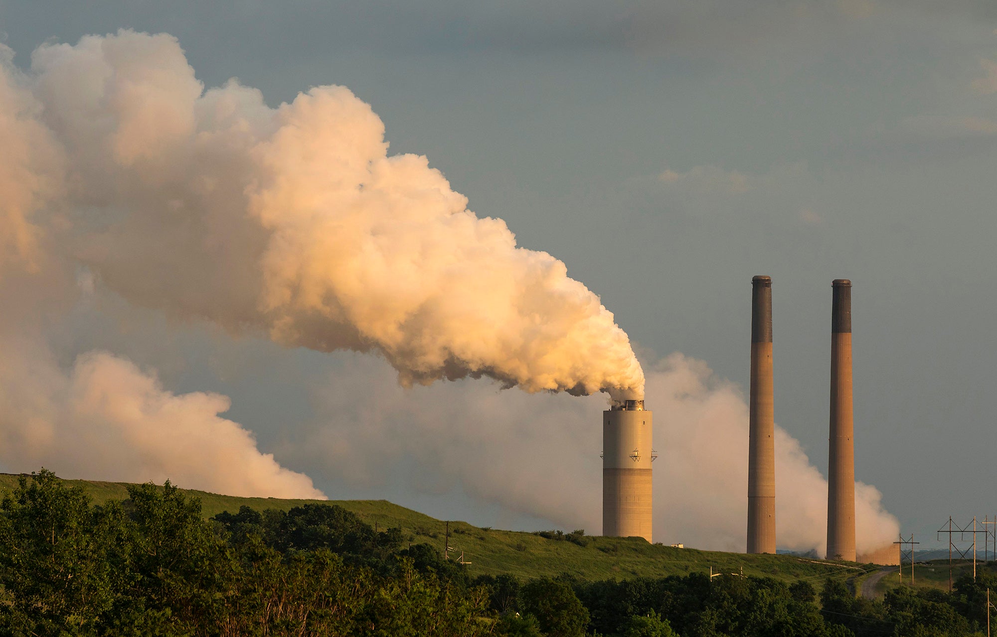 Three smokestacks rise from behind a green hill with one spewing a large amount of white smoke. Smoke it also coming out of two other areas obscured by the hill.