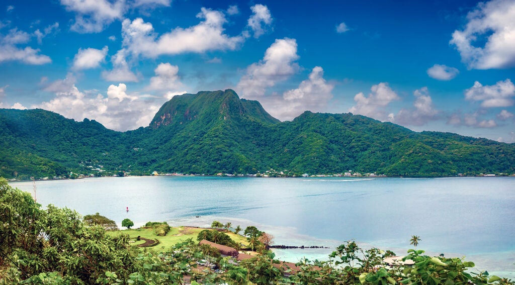 A wide landscape view of a green island wrapping around a large, natural, blue ocean harbor. Small buildings are on the coast with a tall hill or mountain on the far side.