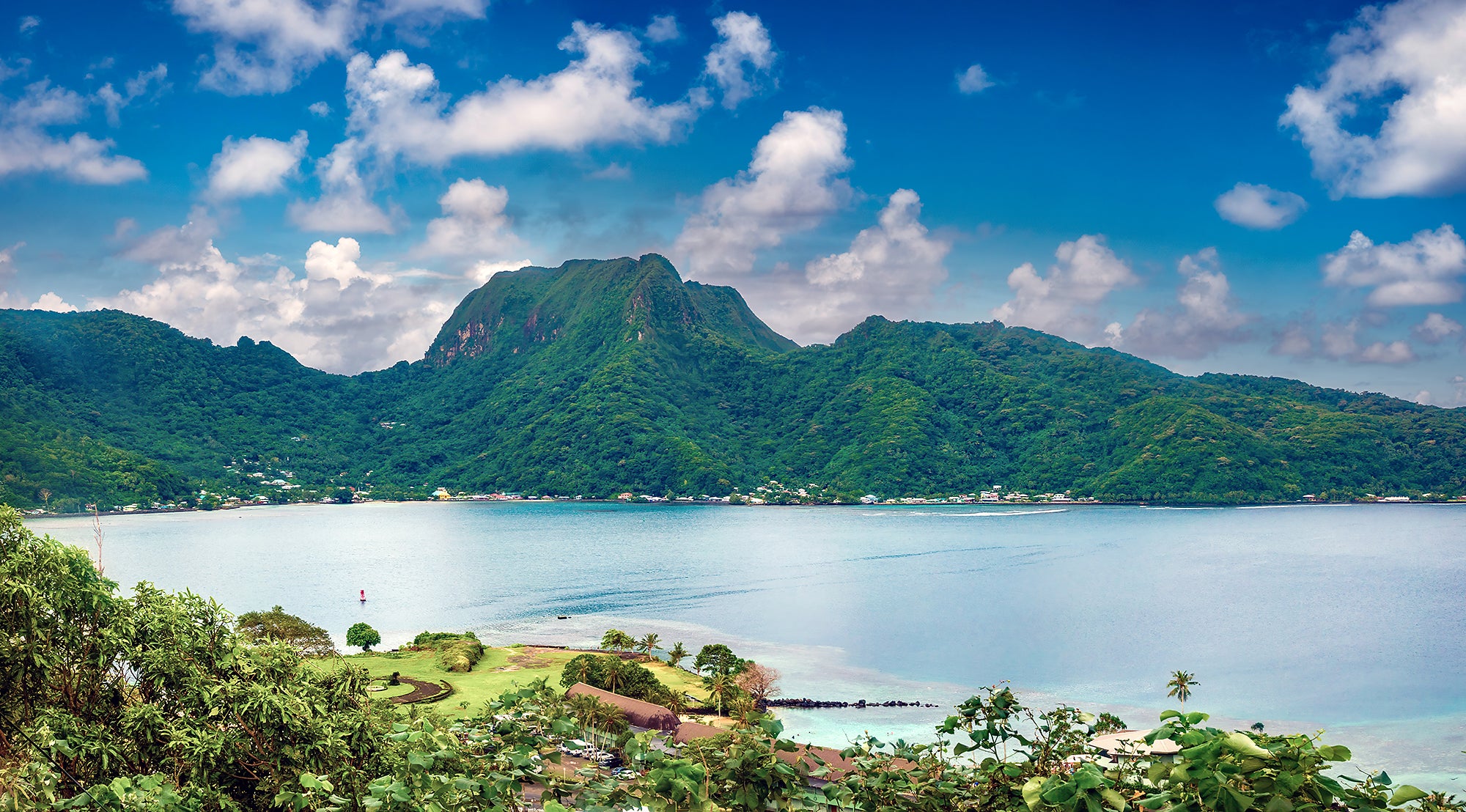 A wide landscape view of a green island wrapping around a large, natural, blue ocean harbor. Small buildings are on the coast with a tall hill or mountain on the far side.