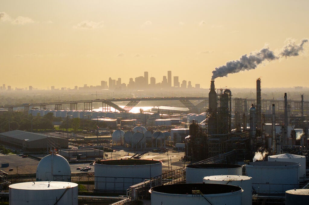 A refinery with storage tanks and exhaust stacks, some releasing vapor, are in the foreground of an aerial photo with the skyline of Houston on the horizon.