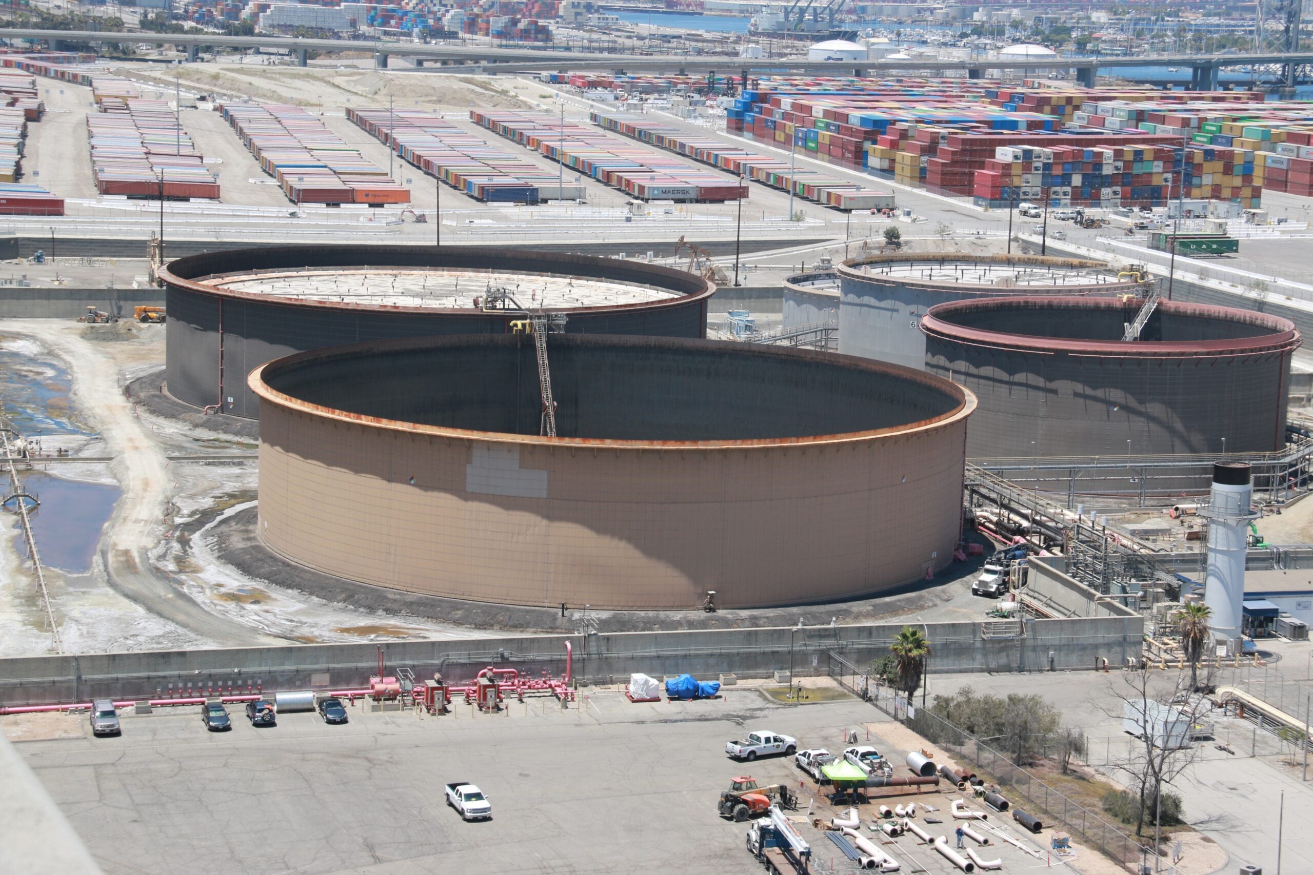 An aerial view of huge oil storage tanks. Two of the tanks in the foreground have large cranes in the middle of them. In the background are many stacked shipping containers in the Port of Long Beach, with ships and the water visible.