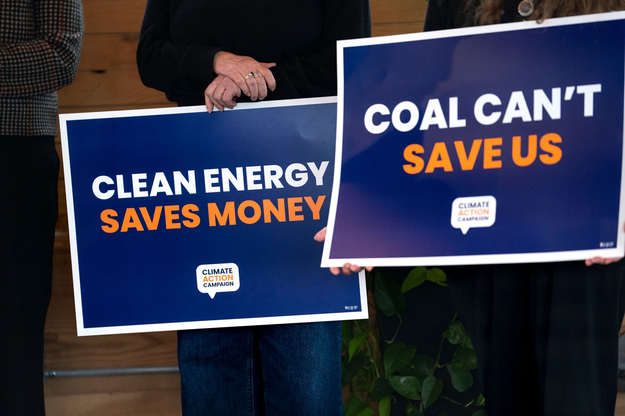 Two large signs -- one reading "Clean Energy Saves Money" and the other, "Coal Can't Save Us" -- are held by two people standing next to each other in an indoor location.
