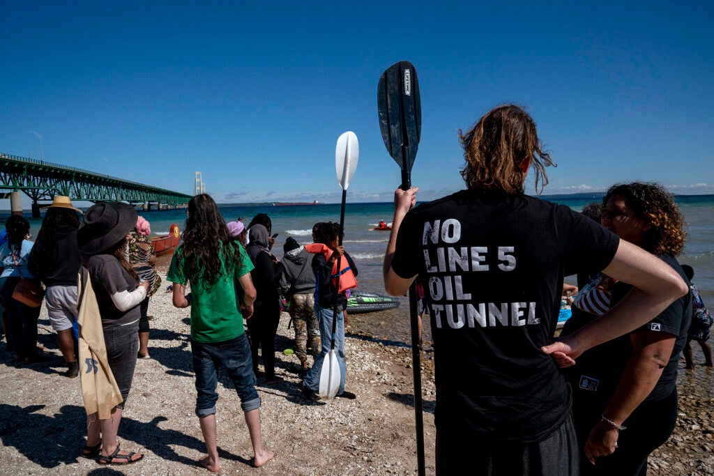 A group of people stand on the bank of a large body of water facing away from the camera, with a large bridge visible on the left of the frame. The shirt on the person on the right reads "NO LINE 5 OIL TUNNEL"