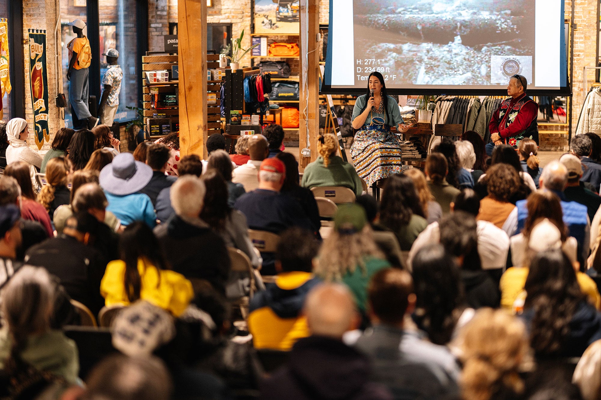 Two people are seated under a large, elevated projector screen inside of a outdoor apparel store, facing a large crowd of seated people. One holds a microphone and is speaking, with a hand outstretched.