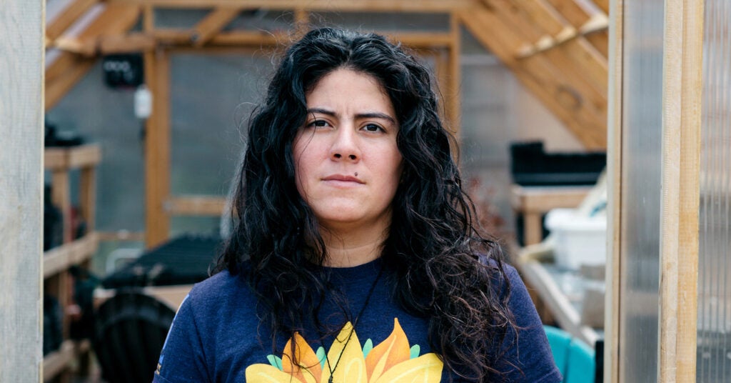 A woman looks directly at the camera with a serious expression standing in the door of a greenhouse.