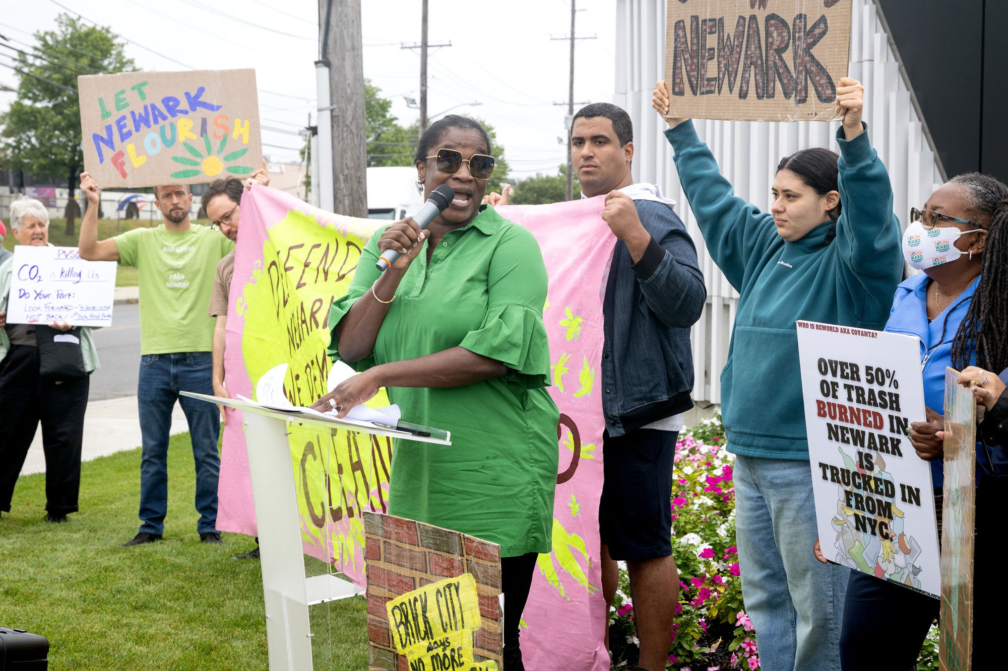 Protestors holding signs that say "Let Newark Flourish," "Over 50% of trash burned in Newark is trucked in from NYC," and more. A woman dressed in bright green and sunglasses, stands in front of a podium with a mic in hand, in front of the crowd of protestors.