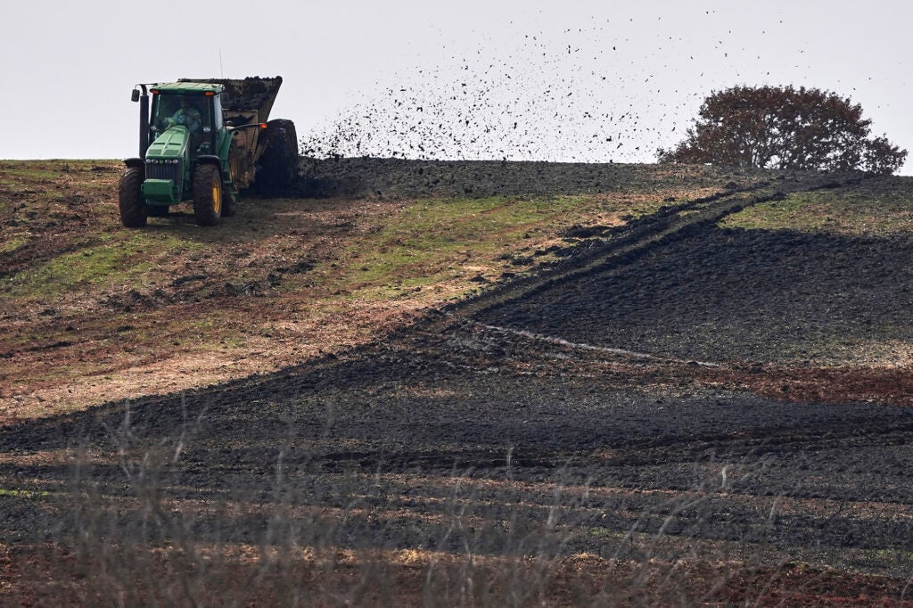 A tractor blows dark, Earth-like material out of a trailer and onto a tilled field.